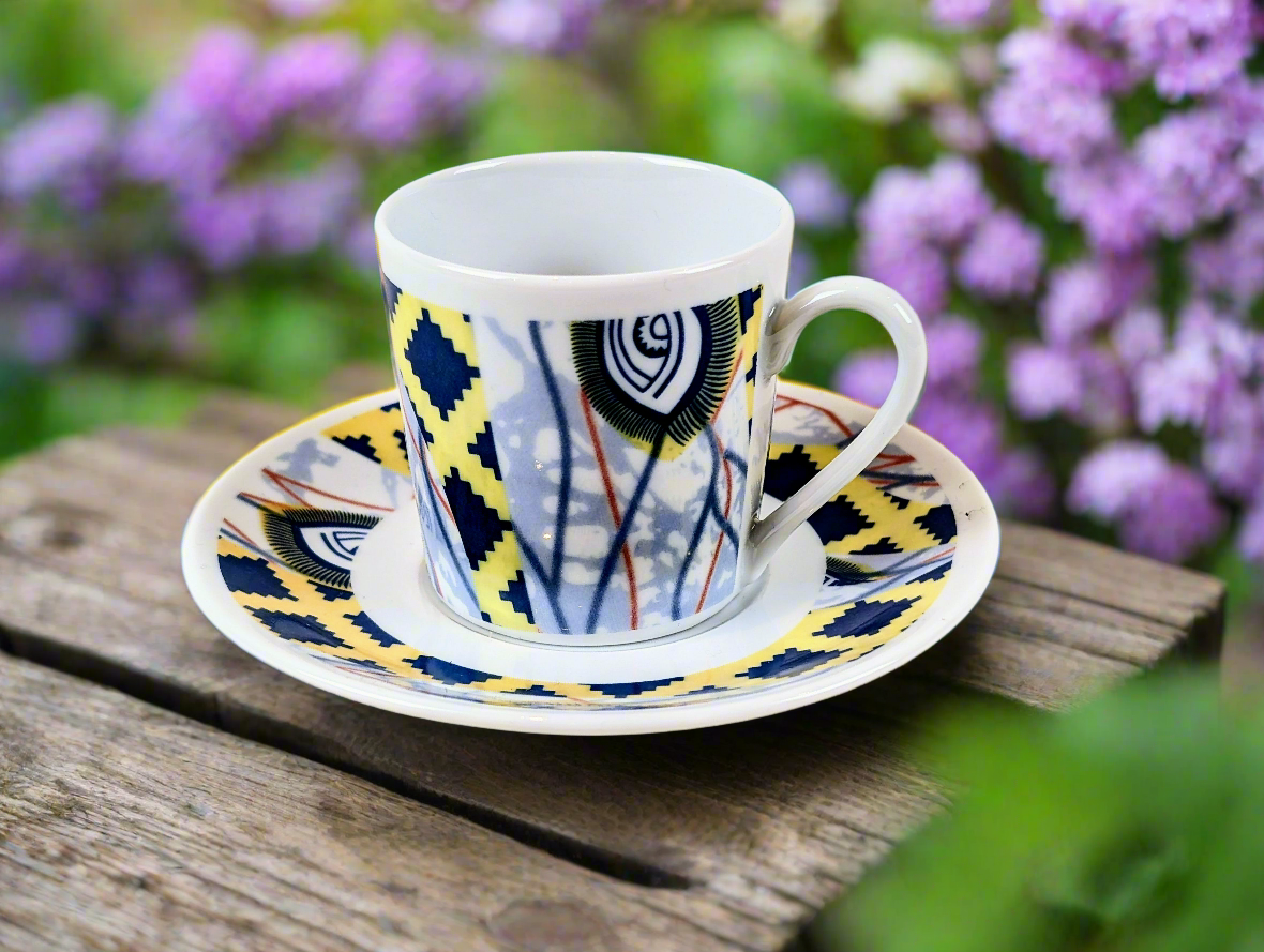 Vue de trois quarts : tasse à café Œil de ma rivale, croisillons jaunes et bleu marine, sur bois devant des fleurs mauves