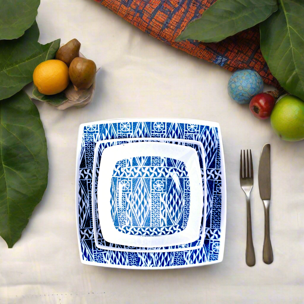Vue zénithale : assiettes carrées Ndop indigo sur toile claire, couverts, kiwis, oranges et feuilles vertes
