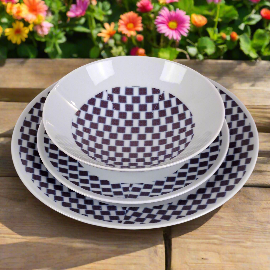 Vue de face légèrement plongeante : bol et assiettes rondes Ludo Kouin à damier violet sur bois, fleurs colorées